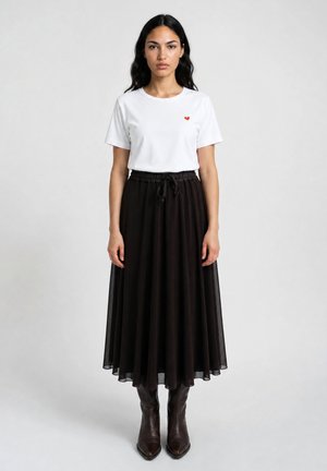 Woman with long dark hair wearing a white t-shirt with a small red heart, a black midi skirt, and dark brown boots, standing against a plain background.