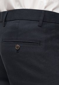 Κοντινή λήψη της πίσω τσέπης σκούρου ναυτικού παντελονιού με κούμπωμα κουμπιού και λευκό πουκάμισο μέσα.