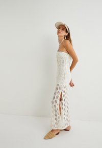 Weißes Häkelkleid mit tailliertem Oberteil und seitlichen Schlitzen, das mit komplizierten Mustern verziert ist. Kombiniert mit einer hellbraunen flachen Sandale.