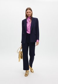Blazer navy con spalline decorate, blusa rosa, pantaloni navy e stivaletti oro lucidi. La modella porta una borsa dorata.