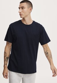 Tricou din bumbac de culoare bleu marin, cu gât rotund, mâneci scurte și un buzunar pe piept. Prezintă o croială lejeră și o textură netedă.