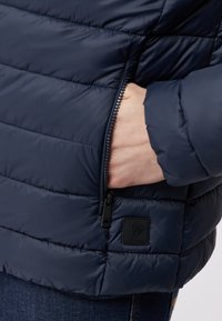 Marineblau gesteppte Jacke mit horizontalen Nähten. Verfügt über eine Reißverschlusstasche und einen strukturierten Patch mit Logo. Glattes, glänzendes Material und taillierte Passform.