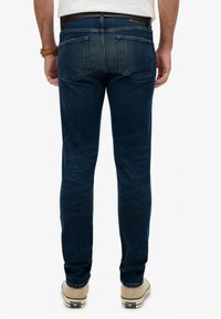 Vaqueros de mezclilla azul oscuro con corte ajustado, costuras visibles y dos bolsillos traseros. Se llevan con zapatillas de color claro.