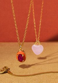 Goldene Halsketten mit einem roten Edelsteinanhänger in einem kreisförmigen orangen Rahmen und einem herzförmigen, rosafarbenen Anhänger. Texturierten Stoffhintergrund.