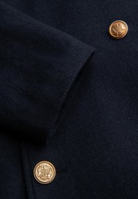 Navy-Wollstoff mit einer weichen Textur, versehen mit zwei goldenen Knöpfen mit filigranen Details auf der Vorderseite.