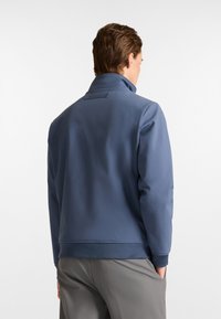 Herren Jacke in Marineblau aus glattem Stoff, mit hohem Kragen, strukturierten Bündchen und einem Logo-Detail auf der Rückseite.