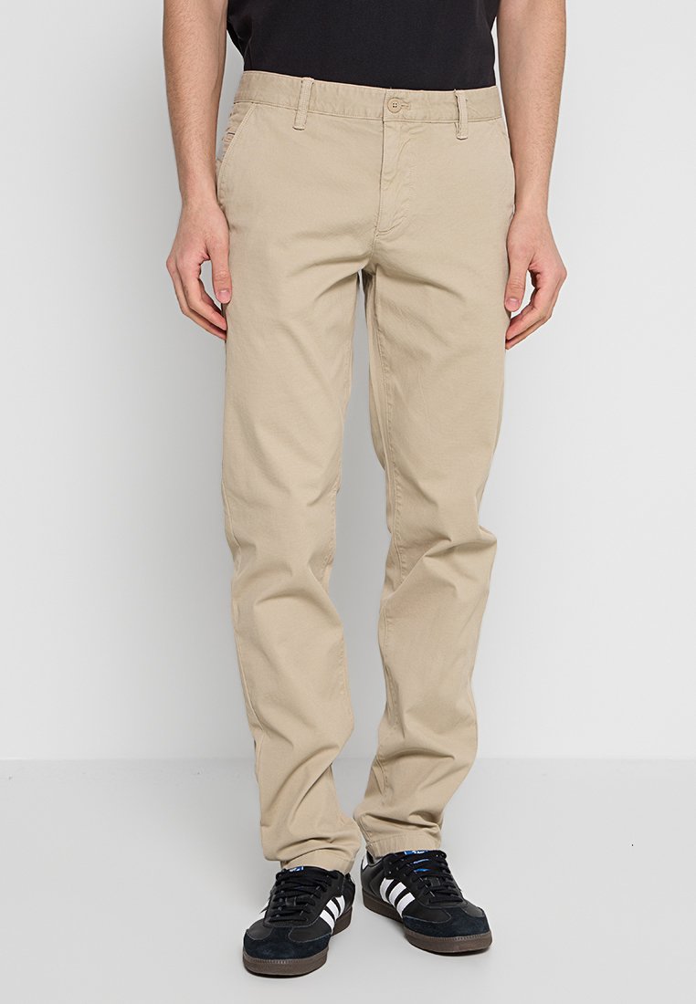 Beżowe chinosy o prostym kroju, wykonane z bawełnianej tkaniny, z zapięciem na guzik i kieszeniami bocznymi. Noszone z czarnymi trampkami.