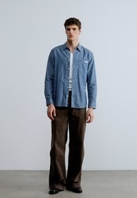 Svetlomodra srajca z gumbi iz tkanine chambray, nošena preko bele majice brez rokavov. Temno rjave široke hlače, črni čevlji. Minimalistična zasnova.