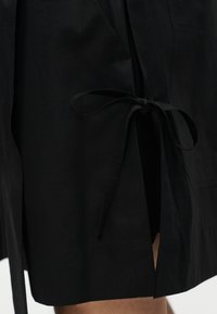 Μαύρη φούστα με πλευρική σχισμή που δένεται με στενό φιόγκο, αποκαλύπτοντας ένα μικρό τμήμα γυμνού ποδιού από κάτω.