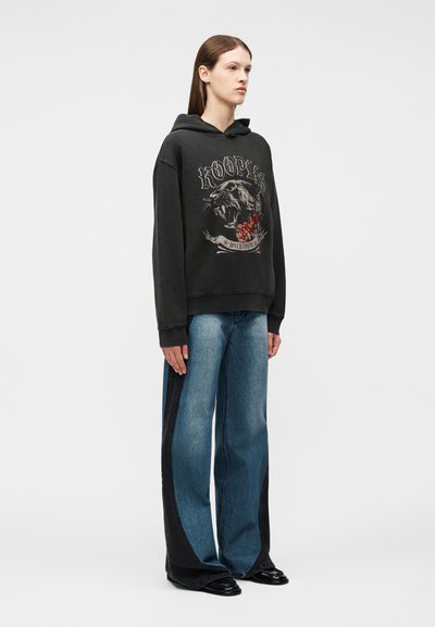 Γυναίκα που φοράει μαύρο φούτερ με κουκούλα με εκτύπωση φοβερής πάνθηρας και λουλουδιών, συνδυασμένο με μπλε και μαύρο τζιν με φαρδιά πόδια και μαύρα παπούτσια.