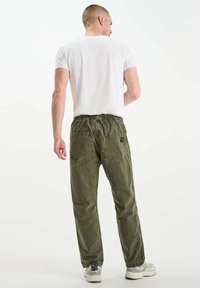 Man in een wit T-shirt, olijfgroene broek met achterzakken en sneakers, staand met zijn rug naar de camera op een effen witte achtergrond.