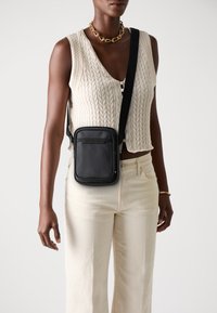 Borsa a tracolla nera di forma rettangolare, con texture liscia e branding sottile. Indossata da un modello con un top a maglia crema e pantaloni chiari.