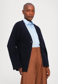 Bald person wearing a ljusblå skjorta med knappar, en navy stor cardigan med svarta knappar, och bruna vida byxor med hög midja.