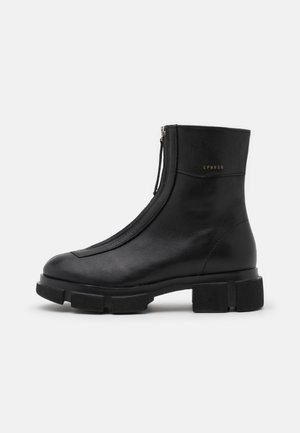 Bottine en cuir noir avec fermeture éclair à l'avant et semelle épaisse crantée, petites initiales de la marque embossées en or sur le côté.
