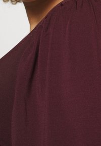 Burgundin yläosa, jossa on pehmeä ja sileä kangas; siinä on laskostetut koristelut hartioissa, rento istuvuus ja syvä kaula-aukko.