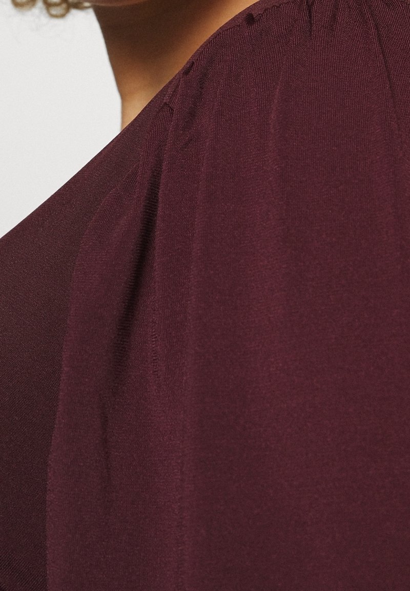 Burgundin yläosa, jossa on pehmeä ja sileä kangas; siinä on laskostetut koristelut hartioissa, rento istuvuus ja syvä kaula-aukko.