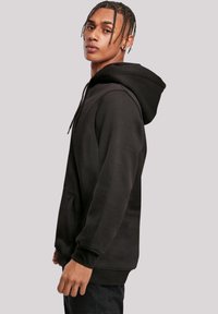 Young man with short braids wearing a black hoodie and black pants, standing sideways against a plain light gray background.
