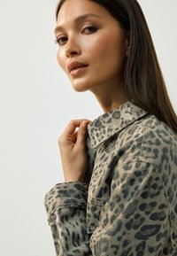Jakna s dezenom životinja s ovratnikom i detaljima na dugmad, u nijansama zemljanih tonova, s teksturiranim materijalom. Model nježno drži ovratnik.