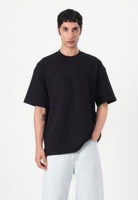 Tricou negru supradimensionat din bumbac ribbed, cu guler rotund și umeri lăsați. Purtați cu blugi albaștri deschiși, modelul are un tatuaj pe braț.