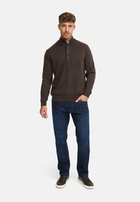 Brun strikket sweater med ribbet krave, tre-knap lukning, parret med mørkeblå jeans og mørke sneakers, stående mod en hvid baggrund.