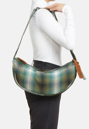 Schultertasche mit grünem, schwarzem und weißem Karomuster, getragen von einer Person mit weißem Langarmshirt und schwarzer Hose, die Hand hält den Riemen nahe der Schulter.