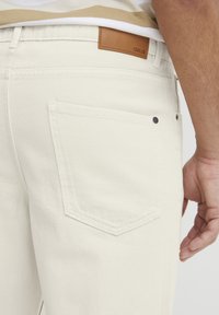Hvite denimjeans har en baklomme med en svart metallnitte og en brun lærmerkelapp på linningen. Synlige sømmer.