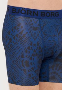 Boxers Björk Borg em azul com um padrão texturizado de estampa animal em preto. Apresenta um cós largo azul com texto do logótipo em preto.