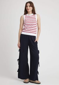 Haut sans manches blanc avec un motif de vagues rouges ; texture tricotée. Pantalons larges navy avec des détails de nouage sur les côtés. Chaussures décontractées avec des brides beiges.