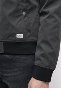 Giacca bomber nera con un materiale esterno a trama liscia, polsini e orlo a coste, e un'etichetta con "#PRODUKT" ricamata nella parte inferiore.