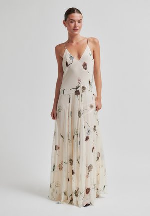 Frau trägt ein ärmelloses, elfenbeinfarbenes Maxikleid mit Blumenmuster, V-Ausschnitt und fließendem Rock, steht vor einem schlichten Hintergrund.