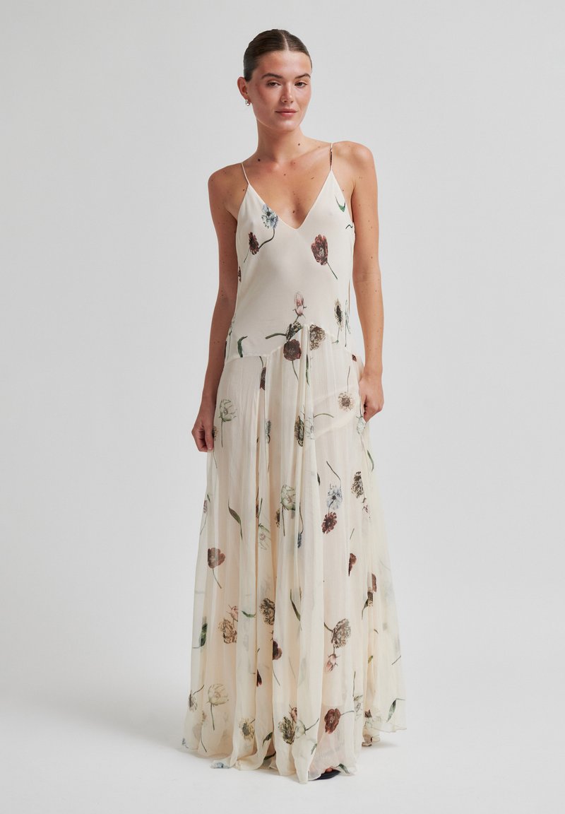 Γυναίκα που φοράει αμάνικο, μακρύ, εκρού φόρεμα με floral σχέδιο, V-λαιμόκοψη και αέρινη φούστα, στέκεται μπροστά σε απλό φόντο.