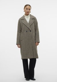 Dulji kaput s dvostrukim kopčanjem u smeškastom i bež uzorku houndstooth, sa velikim reverima i bočnim džepovima, uparen s crnim hlačama i obućom.