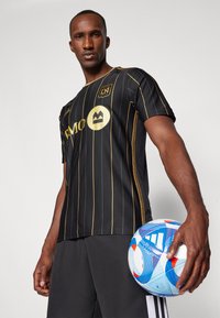 Maglia da calcio a strisce nere e oro con colletto tondo e maniche corte, abbinata a pantaloncini neri. Il giocatore tiene in mano un pallone da calcio blu e bianco.