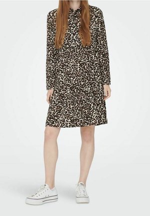 Kobieta w sukience o długości do kolan, z długim rękawem i kołnierzykiem, w panterkę, w białych niskich trampkach, stojąca na jednolitym tle.