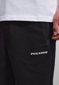 Nahaufnahme einer Person, die schwarze, strukturierte Hosen mit dem "PEGADOR" Logo und einem weißen Hemd trägt, Hand ruht nahe der Tasche.