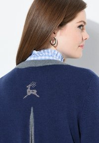 Vrouw met steil bruin haar, die zilveren oorbellen van knopen draagt en een marineblauwe trui met een lichtgrijs rendiernaaldwerkpatroon op de achterkant.