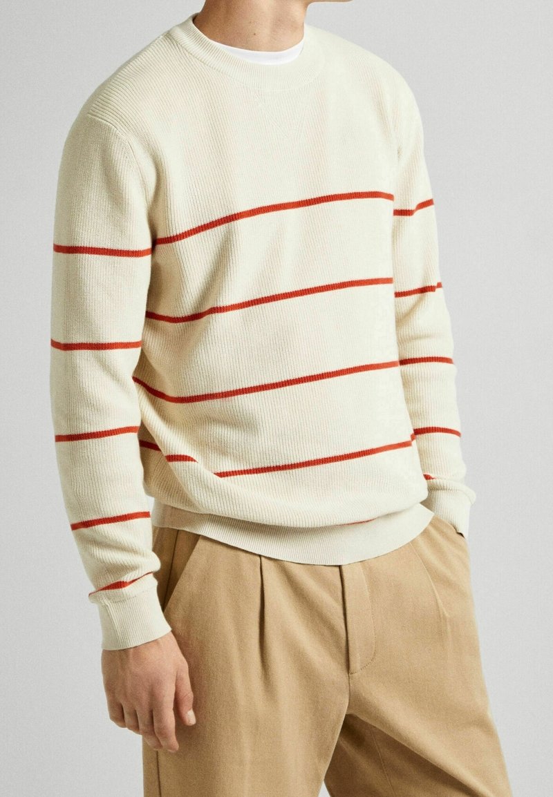 Cremefarbener Pullover mit roten horizontalen Streifen, gerippter Struktur, Rundhalsausschnitt und langen Ärmeln, kombiniert mit hellbraunen, plissierten Hosen.