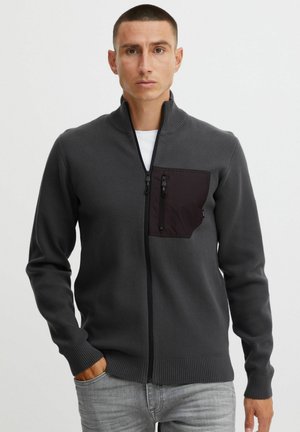 Dunkelgrauer Reißverschluss-Pullover mit hohem Kragen, ausgestattet mit einer schwarzen Brusttasche. Strickstruktur und gerippte Bündchen. Minimalistisches Design.