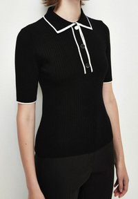 Polo noir côtelé avec col blanc et détails de boutons. Manches courtes avec accents blancs. Coupe ajustée, texture douce.