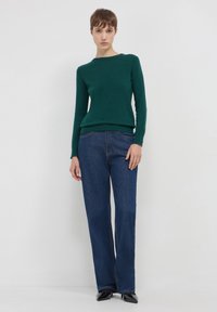 Dunkelgrüner Strickpullover mit Rundhalsausschnitt, kombiniert mit weit geschnittenen, dunkelblauen Jeans und schwarzen, spitzen High Heels. Glatte Textur, bequeme Passform.