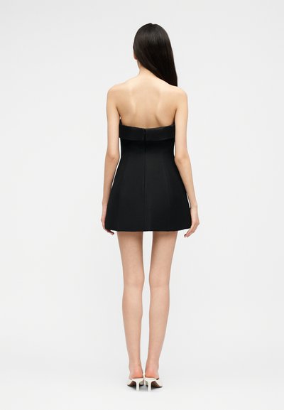 Woman standing facing away wearing a strapless black mini dress and white heeled mules against a plain light background.