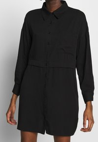 Abito camicia nero con bottoni, maniche lunghe, colletto, tasca sul petto e cucitura in vita, realizzato in un tessuto morbido.