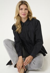 Camisa negra de botón con cuello redondo y mangas abullonadas, combinada con jeans gris claro. Textura suave, corte casual y diseño elegante.
