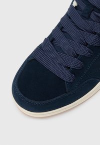 Navy blue suede sneaker with textured fabric, thick navy laces, and cream rubber outsole. Features a rounded toe and stitching accents.
