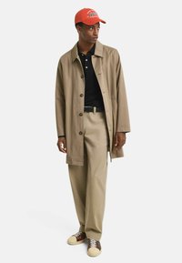 Beige trenchcoat over een zwarte polo, tan broek en bruine sneakers met crèmekleurige details, afgemaakt met een oranje baseballpet.