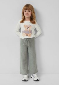 Weißes Langarmshirt mit einem Teddybär-Graphic und dem Wort "SMILE", kombiniert mit hellgrauen, flauschigen weit geschnittenen Hosen und weißen Sneakern.