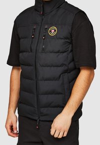 Czarna pikowana kamizelka z wysokim kołnierzem, kieszeniami zapinanymi na zamek oraz naszywką z logo na lewej piersi. Materiał ma gładką teksturę i nowoczesny design.