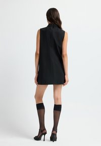 Frau in einem ärmellosen schwarzen Minikleid mit besticktem Kragen, transparenten schwarzen Kniestrümpfen und schwarzen High-Heels, die sich abwendet.