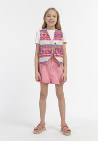 Daugiabriaunis liemenė su geometriniais raštais ant baltos marškinėlių, derinama su rožiniais šortais ir rožiniais espadriliais. Paprastas, kasdienis dizainas.