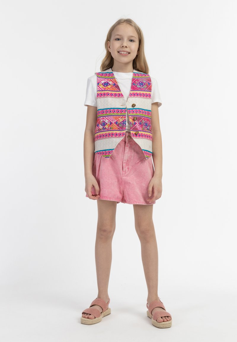 Daugiabriaunis liemenė su geometriniais raštais ant baltos marškinėlių, derinama su rožiniais šortais ir rožiniais espadriliais. Paprastas, kasdienis dizainas.